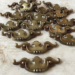 Op de afbeelding: Een groep van 15 antieke messing ladegrepen met een decoratief, sierlijk ontwerp. De grepen hebben een licht aangetast oppervlak en zijn in een verspringend patroon gerangschikt.