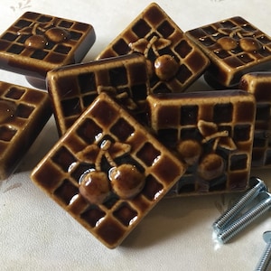 Peut inclure: Ensemble de boutons de placard carrés en céramique marron, avec un motif de gaufre et un design de cerise. Les boutons sont disposés de différentes manières, avec quelques vis visibles. Ces boutons décoratifs sont parfaits pour les armoires de cuisine ou de salle de bain.