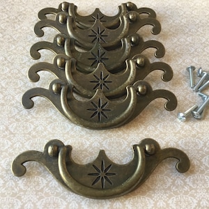 Op de afbeelding: Antieke messingkleurige ladegrepen met een sterrenpatroon. De grepen hebben een gebogen handvat en decoratieve details. Inclusief montageschroeven. Ideaal voor meubelrestauratie of het toevoegen van vintage charme.
