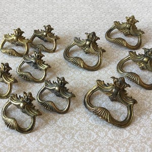 Puede incluir: Un conjunto de diez tiradores de cajón antiguos de latón. Cada tirador presenta un diseño ornamentado con una parte superior en forma de corona y un asa curvada. Aspecto envejecido.