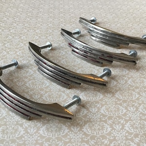May include: Four vintage silver-toned cabinet pulls with a curved, layered design and red accents. Each pull has a screw for mounting. The pulls are arranged on a patterned, off-white surface.