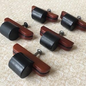 Peut inclure: Cinq poignées de meubles vintage. Chaque poignée présente une poignée cylindrique noire fixée à une base rectangulaire brun-rouge avec une vis argentée. Les poignées sont disposées sur une surface crème à motifs.