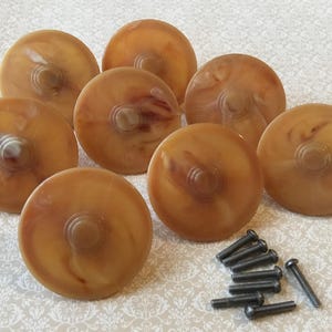 Op de afbeelding: Set van acht vintage-stijl kastknoppen met een gemarmerd amber en bruin ontwerp. Elke knop heeft een centraal, helder, cirkelvormig accent. Inclusief een set zwarte schroeven. Ideaal voor meubelrestauratieprojecten.