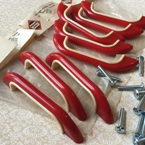 May include: A set of six red and cream-colored cabinet handles with mounting screws. The handles have a curved design and are made of a glossy material. Packaging with text is visible in the background.