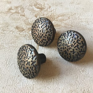 Pode incluir: Três botões de armário de latão antigo com um design circular texturizado. Os botões têm uma borda ligeiramente elevada e um pequeno poste redondo na parte de trás.