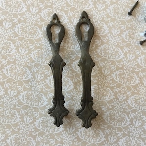 May include: Two antique bronze-colored keyhole escutcheons with ornate detailing, alongside several screws and nails. The escutcheons have a decorative shape and are likely for furniture or cabinetry. The background features a cream-colored floral pattern.
