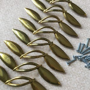 Peut inclure: Ensemble de poignées de tiroir dorées en forme de feuille avec une poignée incurvée, disposées sur une surface à motifs. De petites vis argentées sont éparpillées à proximité, suggérant un ensemble de quincaillerie.