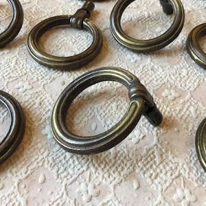 May include: A set of eight antique brass ring pulls, each with a decorative, slightly raised, circular design. The pulls are arranged on a white, floral patterned surface.