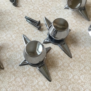 Pode incluir: Uma coleção de objetos decorativos cromados em forma de estrela, brilhantes, com um topo arredondado e três pontas. Dois parafusos também são visíveis. Os objetos estão dispostos sobre uma superfície creme com um padrão floral.