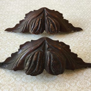May include: Two dark brown, carved wooden corner brackets. Each bracket features a leaf design with a central, textured pod-like element. The brackets are symmetrical and appear to be vintage or antique decorative pieces.