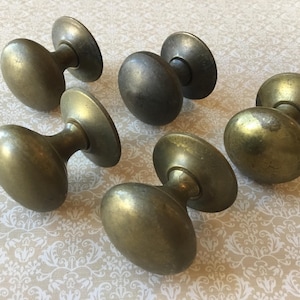 May include: Six antique brass door knobs with an oval shape and circular base. The knobs have a tarnished, aged appearance, suggesting they are vintage or reclaimed. The knobs are made of brass.
