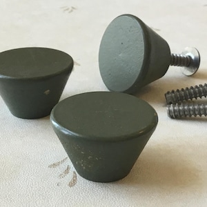 May include: Three olive green cabinet knobs, cone-shaped with silver screws. The knobs have a flat top and a tapered body. Two screws are next to the knobs.