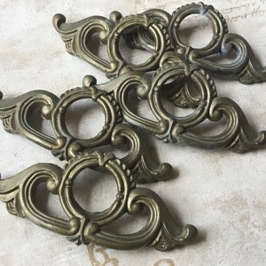 Könnte beinhalten: Drei verzierte Metall-Schubladengriffe mit einem dekorativen Schnörkeldesign. Die Griffe sind messingfarben und haben eine kreisförmige Öffnung in der Mitte.