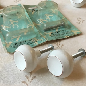 Pode incluir: Dois botões de armário de cerâmica branca com um anel de acento dourado. Os botões são presos a hastes de metal prateado. Os botões são embalados em um cartão azul e branco com o texto "A-527-C1 CERAMOID WHITE-GOLDEN ACCENT" impresso nele.