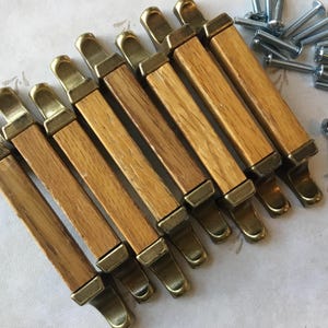 Op de afbeelding: Een set van tien houten kastknoppen met messing accenten. De knoppen zijn rechthoekig en hebben een licht afgeronde rand. De messing accenten bevinden zich aan de boven- en onderkant van de knoppen.