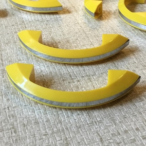 May include: A set of five yellow plastic cabinet door handles with a silver trim. The handles are shaped like a half-circle.