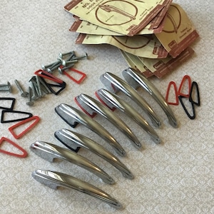 May include: A selection of vintage chrome door handles, screws, and red and black rubber gaskets. Several small brown paper packets with text are also present. The handles have a sleek, curved design.