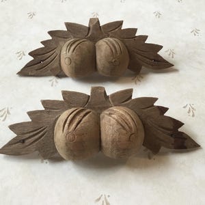 以下が含まれることがあります： 葉のアクセントが付いた果物の木彫り2つ。 各彫刻は、詳細な葉の背景に対して、垂直の溝が付いた2つの丸い果物の形を特徴としています。 彫刻は自然な木の色です。