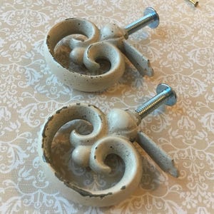 May include: Two off-white, decorative cabinet knobs with a distressed finish. Each knob features a scrolled design and a metal screw for attachment. The knobs are set against a patterned background.
