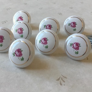 May include: Nine white ceramic cabinet knobs featuring a pink rose design and gold trim. Includes mounting screws. These decorative knobs are perfect for adding a touch of elegance to furniture or cabinets.