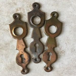 以下が含まれることがあります： 装飾的なデザインが施されたアンティークな真鍮製のキーホールエスカッションが3つ。各エスカッションにはキーホールと、より大きな円形の開口部があります。