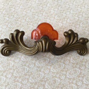 Könnte beinhalten: Verzierter Schubladengriff in Bronzetönen mit einem orangefarbenen, wolkenförmigen Akzent. Der Griff hat ein geschwungenes Design mit dekorativen Verzierungen und einem zentralen orangefarbenen Element, ideal für Möbelrestaurierung.