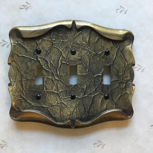 May include: A decorative triple light switch cover with a textured, aged brass finish. The cover has an ornate design with a dark, mottled appearance and black screws. The overall theme is vintage or antique.