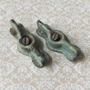 May include: Two antique, cast metal drawer pulls with a weathered, turquoise-green finish. Each pull features a central screw and a decorative, stylized design. The pulls are set against a cream-colored background with a floral pattern.