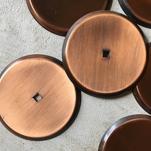 Op de afbeelding: Een set van zes ronde koperen onderzetters met een vierkant gat in het midden. De onderzetters hebben een geborstelde afwerking en zijn in een halve cirkel gerangschikt.