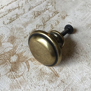 May include: A round, brass cabinet knob with a black screw. The knob has a raised, circular rim around the edge.