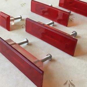 May include: A collection of red rectangular cabinet pulls with clear, rectangular bases and silver screws. One square pull is also present. The pulls are arranged on a light-colored surface with a subtle pattern.
