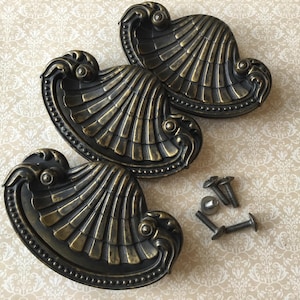 May include: Three antique brass shell-shaped drawer pulls with screws. The pulls feature a detailed, fan-like design with a dark bronze finish and a decorative edge. Suitable for furniture restoration projects.