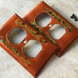 以下が含まれることがあります： 緑と茶色の花柄が施されたオレンジ色のセラミック製コンセントカバー2つ。カバーは薄い茶色の表面の上に置かれています。