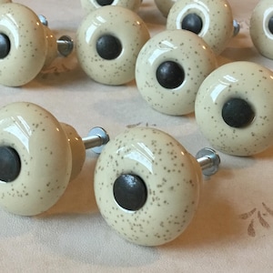 May include: Cream-colored ceramic cabinet knobs with a speckled pattern and a dark center. The knobs have a round shape and are attached to a metal screw. These decorative knobs are suitable for furniture and home decor.