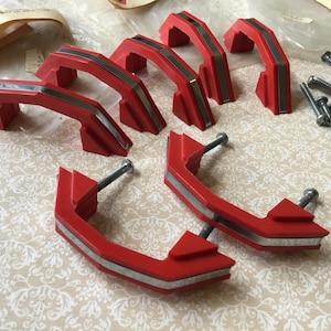 May include: A set of red and silver cabinet handles with screws. The handles have a retro design with a curved shape and a silver accent. The hardware is likely for a kitchen or bathroom, and is ready to be installed.