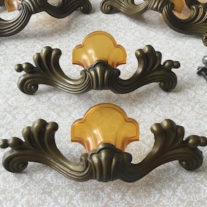 May include: Ornate antique bronze drawer pulls with amber-colored center accents. The hardware features a scrolling design with decorative details. Includes screws for installation. A set of vintage cabinet hardware.