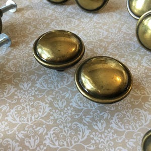 May include: A selection of antique brass cabinet knobs. The knobs are round with a slightly domed surface and a dark rim. The hardware is set against a cream-coloured background with a white floral pattern.