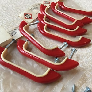 May include: A collection of six vintage red and cream cabinet pulls, each with a mounting screw. The pulls feature a curved design with a cream accent. The hardware is presented on a patterned surface.