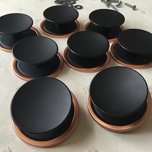 Peut inclure: Huit boutons de cabinet noirs avec une bague en cuivre autour de la base. Les boutons sont ronds et ont un centre surélevé.