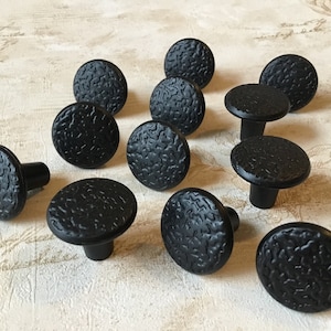 May include: A collection of black cabinet knobs with a textured, hammered appearance. The knobs have a round, flat top and a short, cylindrical stem. The hardware is likely for furniture or cabinetry.