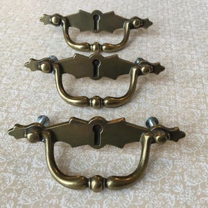 May include: Three antique brass drawer pulls with keyhole details. Each pull has a curved handle and decorative backplate with a keyhole. The pulls are mounted with screws and are set against a patterned background.