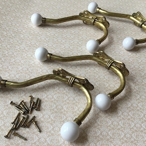 May include: Set of five vintage brass wall hooks with white ceramic ball ends. The hooks have an ornate design and come with mounting screws. The hooks are a decorative and functional addition to any home.