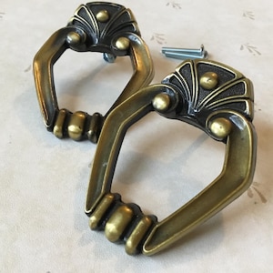 Puede incluir: Dos tiradores de puerta de armario de latón adornado con acabado negro. Los tiradores tienen un diseño decorativo con un centro elevado y una abertura en forma de anillo.