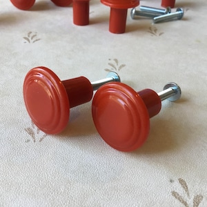 May include: Red cabinet knobs with a rounded, ridged design. The knobs have a cylindrical base and are attached to a metal screw. Several other knobs and screws are visible in the background. These knobs are suitable for furniture.