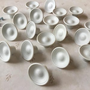 Może przedstawiać: Zbiór 18 białych plastikowych gałek do szafek o gładkiej, zaokrąglonej powierzchni. Gałki są ułożone w losowym wzorze na jasnobeżowej powierzchni.