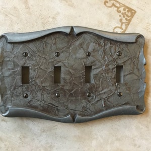 May include: A four-gang light switch plate with a decorative, textured, gray metal finish. The plate has a curved, ornate design and is accented with black screws.