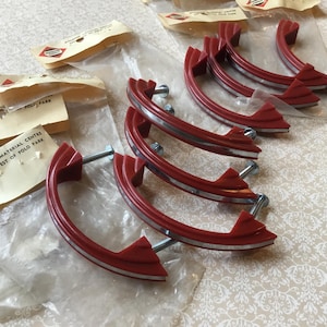 Peut inclure: Ensemble de poignées de placard rouges vintage avec des accents argentés, emballées dans du plastique transparent. Chaque poignée a un design incurvé et est fixée avec un boulon argenté. L'emballage comprend d'anciennes étiquettes en papier avec du texte et un logo.