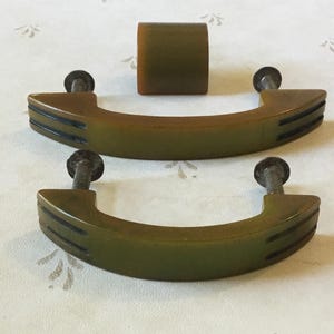 May include: A set of olive green cabinet handles and a knob. The curved handles have three black grooves and metal screws. The knob is cylindrical. The hardware is likely for a drawer or cabinet.