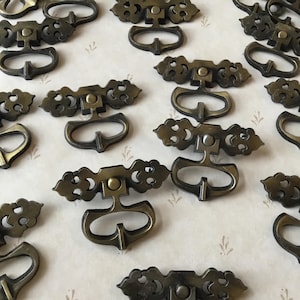 Op de afbeelding: Een verzameling van 14 sierlijke, antieke kastladegrepen. De grepen zijn gemaakt van metaal en hebben een donker bronzen afwerking. Elke greep heeft een decoratief, ingewikkeld ontwerp met een centrale ovale opening.