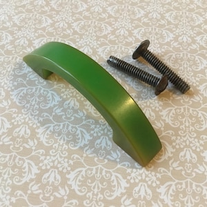 Op de afbeelding: Een groene, gebogen kastgreep met twee schroeven. De greep heeft een afgeronde vorm en een glanzende afwerking. De schroeven zijn van donker metaal met platte koppen. De achtergrond is een crèmekleurige stof met een wit bloemmotief.